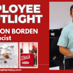 Pharmacist smiling at counter in pharmacy with medication shelves behind, friendly customer service at Mac's Pharmacy, Hamilton Borden pharmacy staff providing healthcare solutions in a welcoming environment.